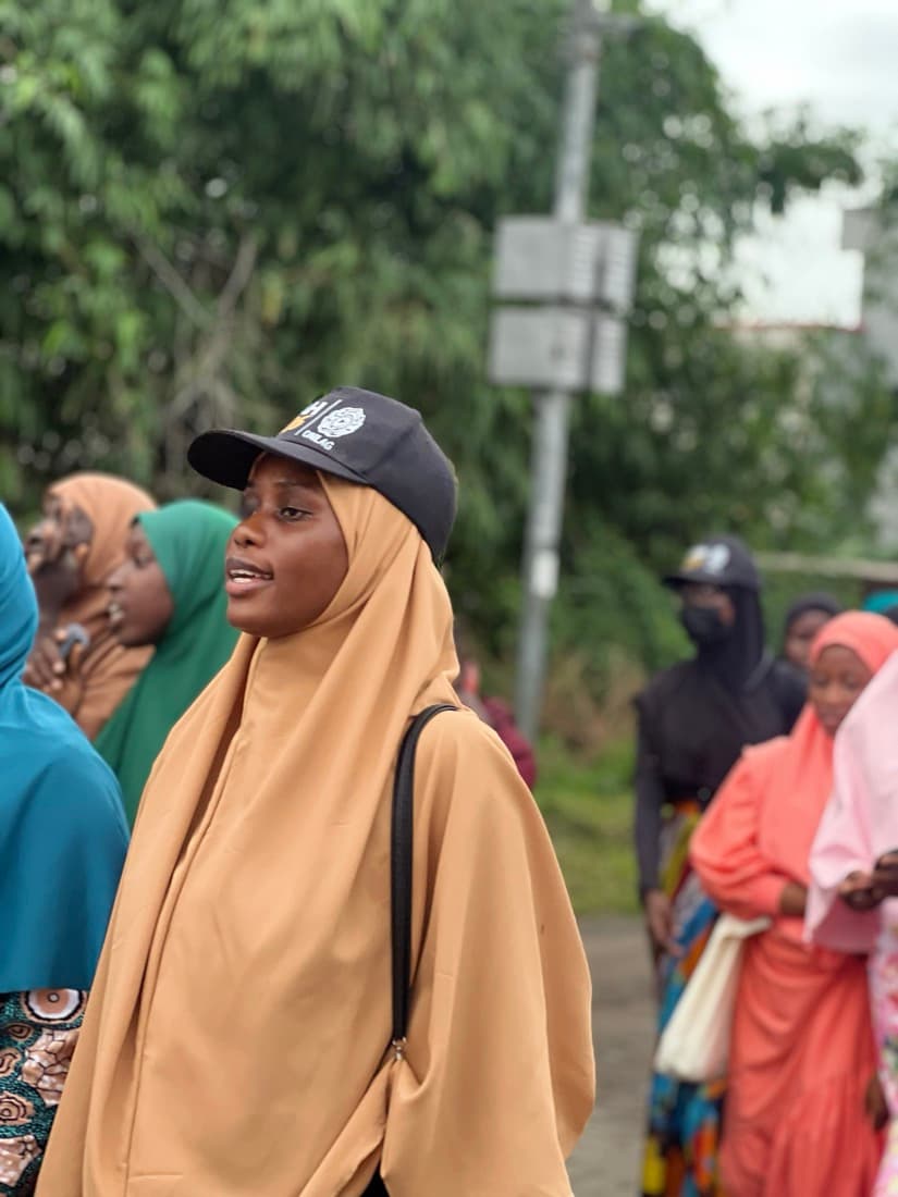 A sister in a mustard hijab and cap among the crowd at the procession