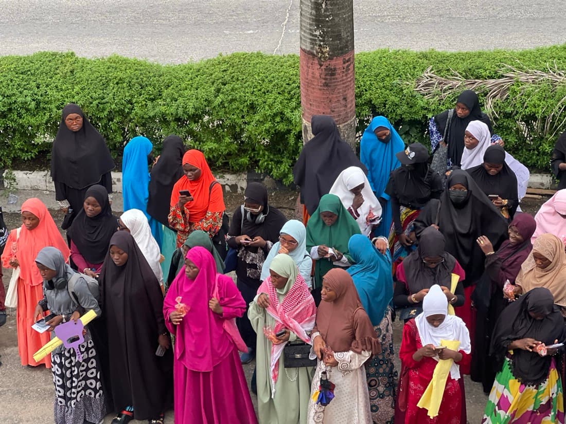 Sisters in colourful hijabs gathered in rows beside a hedge on campus